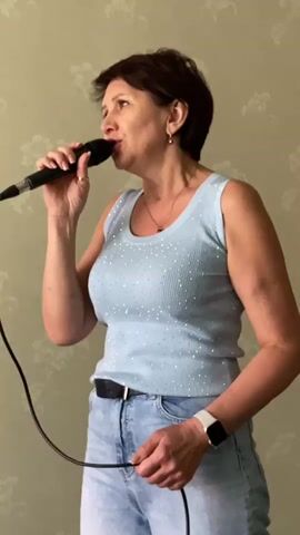 A medium shot captures a woman singing into a microphone. She has short, dark hair and is wearing a light blue, sparkly tank top and blue jeans. She is standing against a patterned wallpaper. Her left hand holds the microphone to her mouth, and her right hand is extended downwards, holding a cable. A smartwatch is visible on her right wrist. The lighting is even, suggesting an indoor setting.