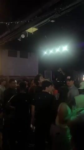 The video shows a crowded indoor venue, likely a bar or nightclub, with dim lighting and string lights overhead. People are standing and socializing, some appear to be dancing. The camera pans across the room, showing different groups of individuals. The atmosphere appears lively, with some people interacting with each other. There are also visible elements of the venue's decor, such as screens and some sort of beverage dispenser. The lighting shifts between red, purple, and blue hues, contributing to a club-like ambiance.