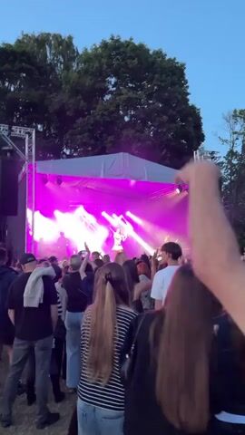 A vertical, outdoor video shows a crowd of people gathered to watch a live music performance on a stage. The stage is illuminated with bright purple and white lights, and there is smoke or haze in the air. A female performer is visible on stage, singing and interacting with the audience. Several band members are also on stage, playing instruments. The crowd is a mix of individuals, many of whom are holding up their phones to record the performance. Trees are visible behind the stage and in the background, suggesting an evening or twilight setting. The overall atmosphere appears to be energetic and celebratory.