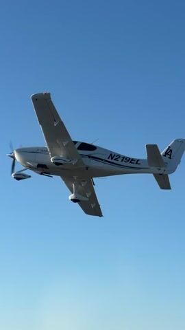 A white airplane with the registration number N279EL is captured in a vertical video, flying through a clear blue sky. The airplane is angled, showing its left side and top, with its propeller spinning. The tail of the airplane has a logo that appears to be the letter "A" inside a diamond shape. In the background, a hazy landscape of rolling hills and scattered vegetation can be seen, suggesting the airplane is flying over a rural or mountainous area. The video transitions from a close-up of the airplane to a wider shot showing its relation to the landscape below.
