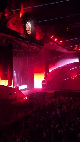 A concert stage is illuminated by vibrant red lights, casting a dramatic glow across the scene. A large, ornate structure hangs above the stage, its details obscured by the darkness and lighting. Silhouetted figures are visible on the stage, likely performers or crew members, with a concentrated beam of white light illuminating a smaller area. Below the stage, a dense crowd of concertgoers can be seen, their faces indistinct in the low light. The overall impression is one of a high-energy, immersive live music event.