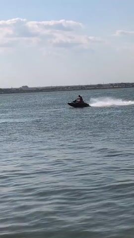 A person is riding a jet ski on a body of water. The jet ski is creating a wake behind it. The water appears to be calm with some small ripples. There is a distant shoreline visible with some structures on it. The sky is blue with some scattered clouds. There are reeds or tall grass in the foreground.