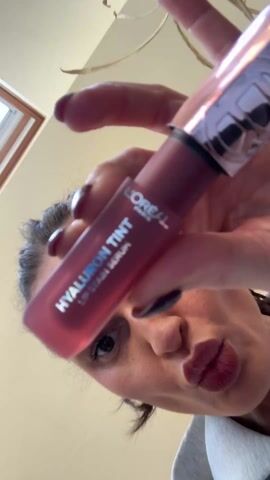 A woman with dark hair pulled back from her face is holding a tube of L'Oreal Paris Hyaluron Tint Lip Stain Serum and a mirror toward the camera. She smiles, opens the lip gloss, and begins to apply it to her lips. She is indoors with a window and lighting fixture visible in the background.