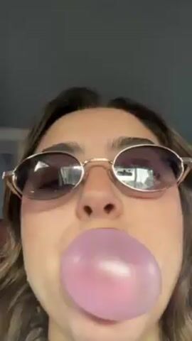 A young woman with fair skin and brown hair, wearing small, gold-rimmed sunglasses, is seen from a low angle, filling her mouth with pink bubblegum. The bubble pops, and the gum is contained within her mouth. The environment appears to be inside a car, as suggested by the interior background.
