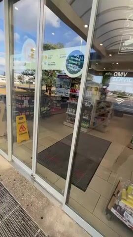 The video begins by showing the exterior of a convenience store, specifically the glass doors. Through the glass, the interior of the store is visible, displaying shelves stocked with various products like drinks and snacks. The store appears to be a "Viva Billa" branded store. The camera then moves inside the store, panning across the aisles. The aisles are lined with shelves filled with an assortment of items, predominantly food and beverages. Products are neatly arranged on the shelves, with labels and packaging clearly visible. The store is well-lit with overhead lighting, and the floor is tiled. There are no visible customers or staff members in the immediate vicinity during the recorded segment.