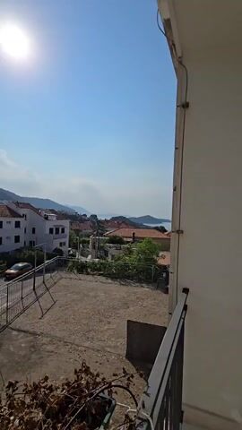 The video pans across a residential area with buildings and a view of the sea. The sun is bright in the sky. There are several cars parked along the street. A metal fence encloses a patch of bare ground. The foreground shows a railing and some dry vegetation.