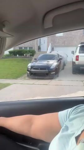 From inside a vehicle, a black Nissan GT-R parked in a driveway is visible. The house at 245 is a brick two-story with white trim. A beige SUV is also in the driveway. The camera moves forward and stops. A person’s arm wearing a light blue t-shirt is visible on the left side of the frame.