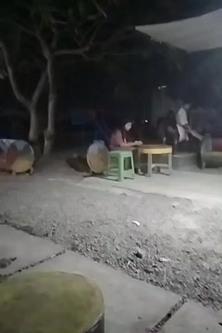 The video shows an outdoor scene at night. In the foreground, there are concrete slabs and gravel. To the left, a round object with yellow and red bands is visible near a tree. In the midground, a person is sitting at a round table with a yellow surface, next to a green stool. Behind them, a food stall is illuminated by bright lights. The stall has a red facade with the word "FOOD" at the top and "SAE" in the middle. Other people are visible in the background, some sitting on benches or standing. Motorcycles are parked in front of another building in the distance, which is also lit. The overall environment appears to be a casual outdoor eating area.