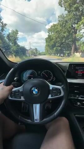 A first-person perspective video shows a person driving a BMW car. The driver's hands are on the steering wheel, which has the BMW logo in the center. The dashboard displays the car's speed and other information. The car is driving on a paved road through a rural area with houses and trees lining the sides. The sky is cloudy with patches of blue. The video appears to be a short clip, possibly taken while on a road trip or commute.