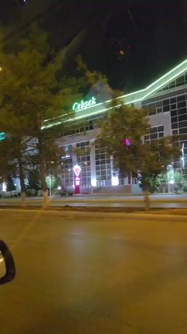 The video shows a nighttime drive through a city street in Ashgabat, Turkmenistan. The scene is dominated by modern, well-lit buildings with illuminated signs and decorative lighting. 

On the right side of the street, a building with the sign "Cebsek" in green neon lettering is visible. It has large windows and is illuminated from the outside. Next to it, another building with a red logo and what appears to be a neon sign of an ice cream cone is illuminated.

Further down the street, on the left, is a white building with bright blue neon lighting outlining its edges. The sign on this building reads "Tajir" in blue neon letters. This building also features large windows and is brightly lit.

The street itself is paved and appears wet, reflecting the lights of the buildings and streetlights. There are trees and other greenery lining the sidewalk in front of the buildings, also illuminated by the surrounding lights. A streetlamp with a warm yellow light is visible in the upper left corner of the frame. The overall impression is that of a well-developed urban area at night, with an emphasis on modern architecture and decorative lighting. The weather appears to be clear, given the darkness and the reflections on the wet road. The activity taking place is a vehicle traveling along the street.