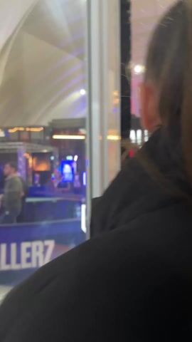 The video shows a person with brown hair pulled back in a ponytail. The person is wearing a black jacket and walking through a large indoor space, possibly a convention center or event hall. The space is brightly lit with numerous overhead lights and colorful digital displays. In the background, there are booths and signage that seem related to sports or entertainment. The person's movement is captured from behind, giving a slightly shaky and blurred perspective as they walk through the event.
