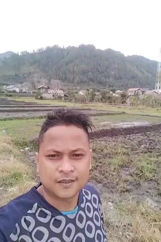 A man in a blue and grey patterned shirt is taking a selfie outdoors. He has short, dark hair and is smiling at the camera. In the background, there are fields with some muddy patches and dry grass, as well as a few houses and a tall communication tower. Hills covered in trees rise in the distance under a cloudy sky.