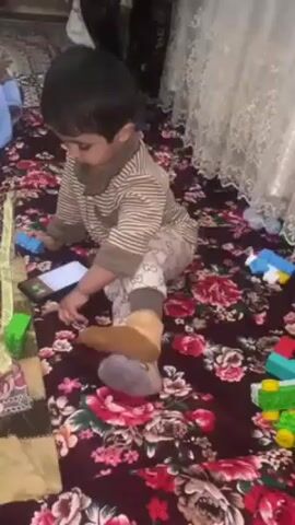 A young child sits on a maroon floral patterned rug, wearing a brown and white striped pajama top and patterned pants, and tan socks with a grey pompom detail. The child is surrounded by colorful plastic toy blocks. A smartphone lies on the rug near the child's feet. The child looks towards the phone then turns to look at the camera. White lace curtains are visible in the background.