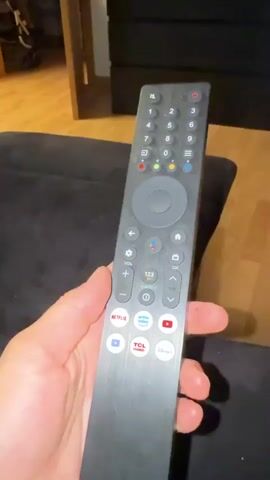 A person's hand is holding a black remote control. The remote has various buttons, including a number pad, directional controls, and shortcut buttons for streaming services like Netflix, Prime Video, YouTube, TCL, and Disney+. The person's thumb is pressing one of the buttons on the remote. The background shows a couch and a doorway leading to a room with wooden flooring.