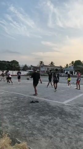 A group of eight young people are playing volleyball on an outdoor court. The court appears to be made of concrete, with a net set up across the middle. The sky is cloudy and it appears to be late afternoon or early evening. The players are dressed in casual clothing, some wearing jerseys with numbers on them. The person in the foreground is wearing a black t-shirt and shorts and is holding a volleyball. The other players are scattered across the court, with some in positions to receive a serve or spike. The background shows a few buildings and trees, suggesting a residential or semi-rural area. The overall atmosphere is one of casual recreation and friendly competition.