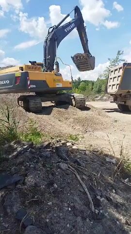 This media file captures a scene at a construction or demolition site in Rozdil, Ukraine. The primary subjects are a large yellow Volvo EC250DL excavator and a gray dump truck. The excavator, with its arm extended, is actively dumping soil or debris into the back of the dump truck. The cab of the excavator is visible, and a person appears to be operating it, though their features are not clearly discernible.

The setting is an outdoor, industrial area characterized by dirt, piles of excavated material, and sparse vegetation. In the background, a line of trees suggests a rural or semi-rural environment. The sky is a bright blue with scattered white clouds, indicating fair weather and likely daytime. The lighting suggests it's a sunny day, possibly late morning or early afternoon.

Visible text includes "VOLVO" prominently displayed on the excavator's boom, and "EC250DL" on the side of the excavator. There are also smaller logos on the excavator, including one that appears to be "SMIS." The dump truck has a textured surface on its rear cargo area, which is typical for such vehicles.

The overall activity is the loading of material onto the dump truck by the excavator, a common process in earthmoving and construction operations. There are no explicit emotional cues conveyed, the scene is focused on the industrial task at hand.