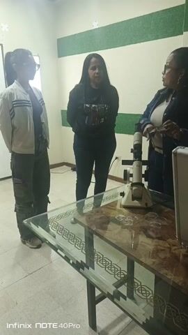 Three women stand around a glass table with a lensometer on it. The woman on the right is holding a pair of glasses and appears to be explaining something about them. The other two women are listening. The table has some extra glasses and a white case on it. The room has white walls with a horizontal green stripe. One of the walls also has a window with sunlight streaming in. The text “Infinix NOTE 40 Pro” is at the bottom of the video.