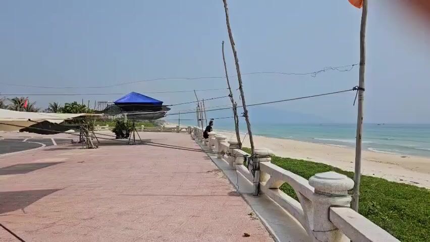 The video captures a serene coastal view in Bac Cam Ranh, Vietnam, on a bright, clear day. The foreground features a wide, reddish-tiled paved area, bordered by a white concrete railing with decorative balusters. Beyond the railing, a strip of green grass precedes a sandy beach stretching along the coastline.

The calm blue ocean is visible in the background, with gentle waves breaking on the shore. Distant mountains or hills are faintly silhouetted against the clear, light blue sky. A single person stands in the mid-ground near the white railing, looking out towards the ocean.

Further back on the left side of the paved area, a blue tent or tarp covers what appears to be structures or boats, with other covered items nearby. Several tall, slender wooden poles with wires strung between them line the edge of the paved area, and a small orange flag is visible on the pole to the far right. The scene conveys a sense of tranquility under abundant sunlight. No visible text is present in the video.