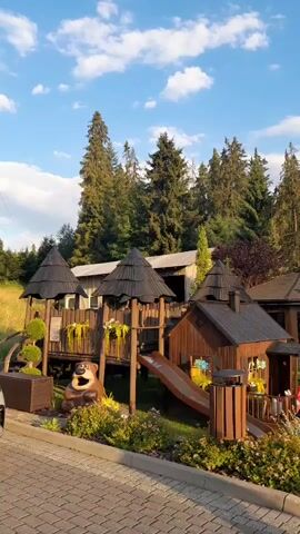 A wooden playground structure with a slide is situated in a natural environment with trees and blue sky. A brown bear statue is placed in front of the playground. There are plants and flowers around the playground.