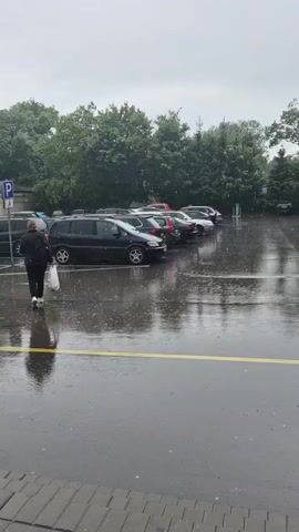 A person walks across a wet, paved parking lot. It is raining, with visible raindrops hitting the puddles on the ground. Several cars are parked in the lot, and a line of trees is visible in the background. The sky is overcast.
