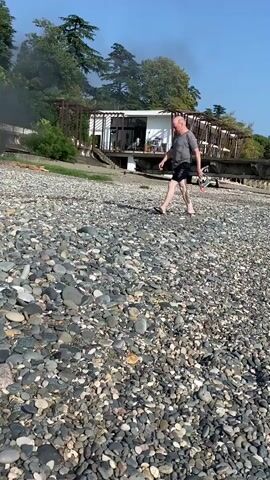 A man walks on a rocky beach. He is wearing a grey t-shirt and black shorts. He is carrying a bottle in his right hand. Behind him, there is a building with wooden accents and a fire burning. Trees are visible on a hill behind the building. The sky is blue.