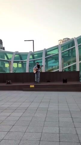 A man sings and plays an acoustic guitar while standing on a small stage. The stage is outdoors and paved with gray tiles. There is a yellow warning sign on the front of the stage with text in Korean. Behind the stage is a modern-looking pedestrian bridge with a green-tinted glass roof. Tall buildings are visible in the background. The man is wearing a light blue shirt, jeans, and brown shoes. He is facing forward and playing the guitar with his right hand and holding the neck of the guitar with his left hand. There is a microphone stand in front of him with a tablet attached. The tablet likely displays the lyrics to the song.  Two small black speakers sit on the stage, one on either side of the man. The sky is a pale blue color, suggesting it is either early morning or late afternoon.