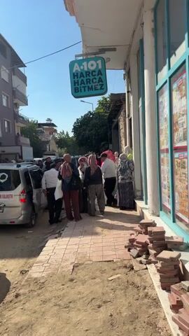 A group of people are gathered outside a store. A sign above the store reads "A-101 HARCA HARCA BITMEZ". Some people are wearing headscarves. A car is parked to the left of the group. The ground in front of the store appears to be under construction, with loose bricks visible. The sky is blue and the sun is shining.