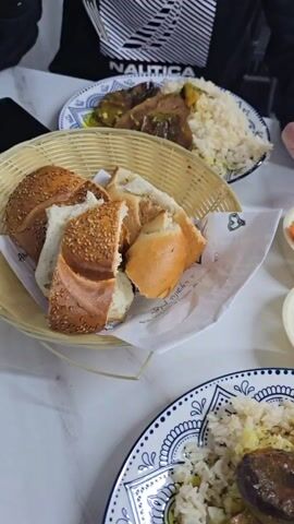 The video showcases a dining table arrangement with a spread of food. The table is white marble. There are plates with rice and meat, accompanied by various side dishes in small white bowls, and a basket filled with bread. People are seated around the table. A person is seen picking up a napkin.