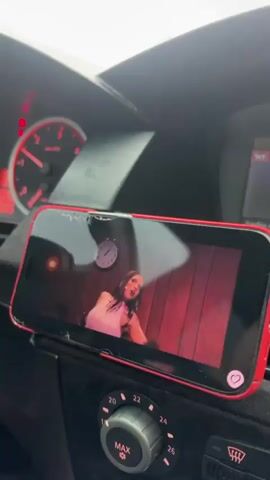 From inside a car, a phone in a red case sits on the dashboard, displaying a video. The video shows a woman singing on stage, wearing a pink top and skirt. There are other people visible on stage with her. The car's dashboard includes climate control knobs and a partially visible speedometer with red markings.