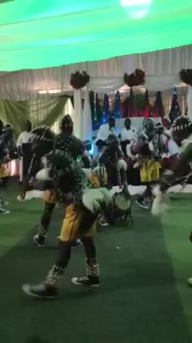 A group of people wearing traditional African costumes perform a dance inside a tent. The tent is decorated with white curtains, green and white fabric, and green carpet. Some people are sitting on chairs and watching the performance. The dancers are wearing costumes with black and white patterns, yellow accents, and sneakers. Some of them are carrying drums. The lighting is a combination of white and green.