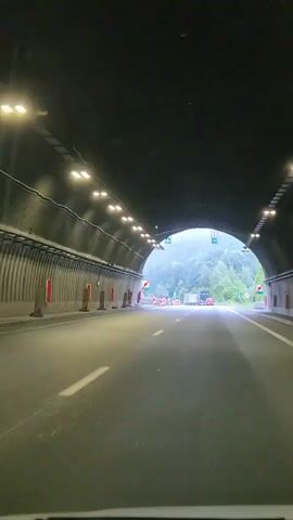 The video captures a first-person perspective of driving through a tunnel and then emerging onto a highway in a mountainous, foggy environment.

The scene begins inside a tunnel, characterized by its dark, arched ceiling illuminated by rows of bright lights. The tunnel walls are ribbed, and traffic cones and temporary barriers are visible on the side of the road. Green directional arrows are illuminated above the roadway, indicating traffic flow.

The vehicle then exits the tunnel into a bright, overcast day with heavy fog or mist obscuring the surrounding mountains. The highway is paved and has clear lane markings. Red and white striped barriers line the sides of the road, and traffic signs, including directional arrows and what appears to be a road closure or lane restriction sign (with a red diagonal line through a symbol), are visible. Other vehicles, including trucks and cars, are on the road ahead. The landscape is densely forested with lush green trees.

The weather appears to be foggy or misty, creating a somewhat subdued and atmospheric mood. The location is identified as Vrachesh, Bulgaria, suggesting a mountainous region within the country. No people are explicitly visible in the footage. The overall activity is straightforward: driving. The time of day is not definitively clear due to the overcast conditions, but it appears to be daytime.