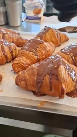 The video shows a tray of freshly baked croissants being decorated with chocolate and sprinkles. The croissants are golden brown and flaky, arranged on a sheet of white parchment paper on a metal tray. A gloved hand sprinkles colorful sugar decorations on some of the chocolate-covered croissants. The background includes kitchen equipment like a metal pitcher and a bag of an unidentified baking ingredient.