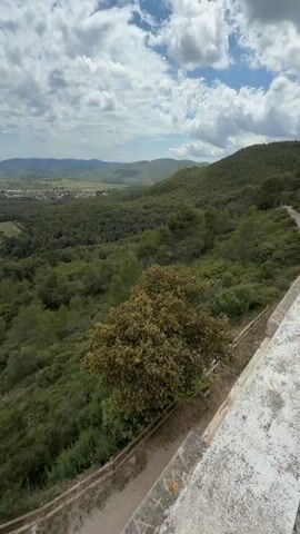 The video shows a panoramic view from a high vantage point, likely a tower or hilltop, overlooking a lush, green landscape dotted with trees and foliage. In the distance, there is a town nestled among the rolling hills. The sky is partially cloudy, with patches of blue breaking through. The camera pans slowly, revealing the varied terrain and the extent of the scenery. The foreground includes parts of the structure from which the video is taken, showing a weathered stone surface and a simple railing.