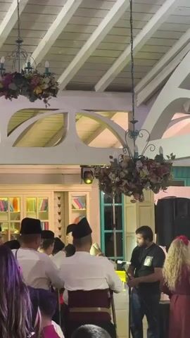 A group of men wearing white shirts and black hats are gathered in a room with a white ceiling and chandeliers. One man is playing a bagpipe, while another man stands next to him holding a microphone. A crowd of people are watching the performance. There are bookshelves and a speaker in the background.