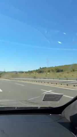 This is a first-person perspective video taken from inside a vehicle driving on a highway. The sky is clear and blue. On the left side of the highway, there is a grassy hill with some trees and a few cows grazing. A guardrail runs along the side of the road. In the distance, there is a large power line tower. The highway itself is paved with visible lane markings. There are other vehicles on the road, including cars. The video captures the view through the windshield, which shows some smudges and a small sticker attached to the glass. Further down the road, a bridge or overpass is visible. The dashboard of the vehicle is partially visible in the foreground.