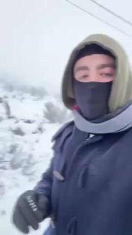 A person dressed in winter clothing stands in a snowy landscape. They point at a snowman made of three snowballs. The snowman has twig arms, a carrot nose, and button eyes. On top of the snowman's head is a red and white can of Super Bock beer. The weather is foggy, and the background is blurry.