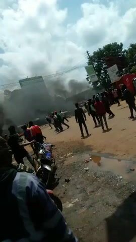 The video depicts a chaotic scene in a town or city. Thick smoke billows in the background, suggesting a fire or some other significant event. Several people are milling around in the street, some walking, some standing, and some engaging in what appears to be a physical altercation. In the center of the frame, a group of individuals is seen fighting, with shoving and pushing visible. The presence of several motorcycles parked nearby suggests that the area is a street or intersection. The surroundings are somewhat rugged, with scattered debris on the ground and puddles of water. Advertisements and signage are seen on the sides of buildings. The overall impression is one of unrest and disorder.