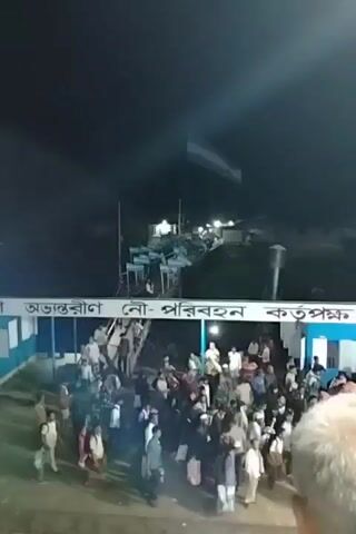 The video captures a bustling night scene, likely in Bangladesh, given the visible Bengali text. A large crowd of people is gathered, some walking along an illuminated walkway or pier. The area is lit by artificial lights, piercing through the darkness of the night sky.

A prominent feature is a large flag, with distinct light blue and white horizontal stripes, resembling the flag of Argentina, waving in the upper part of the frame. Numerous smaller flags, identical in design to the large one, line the entire length of the walkway, creating a festive atmosphere.

In the foreground, a white banner with Bengali text is visible. The text reads "নৌ- পরিবহন," which translates to "Water Transport" or "Naval Transport." This suggests the location is likely a ferry terminal, a port, or a similar facility related to water transportation. People are seen moving in various directions, indicating a lively gathering or transit point.