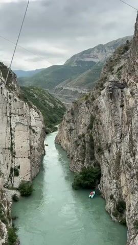 A wide shot shows a turquoise river flowing between steep, rocky canyon walls. Two small boats, one yellow and one white with green accents, are visible on the water. The white boat is closer to the foreground and appears to be moving downstream. The yellow boat is further upriver, also moving downstream. The canyon walls are a mix of light gray and textured brown rock, with patches of green vegetation growing on ledges and at the base. In the background, more distant mountains are visible, with a road winding its way up one of the slopes. The sky is overcast with gray clouds. The video appears to be filmed from a bridge or elevated position, as evidenced by wires or cables visible at the top of the frame.