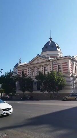The video shows a view of a street in a city on a clear, sunny day. In the foreground, there are several cars, including a yellow taxi on the left and a white hatchback on the right. The cars are driving on a paved road. In the background, there are several buildings, including a large building with a dome and columns, which appears to be a historical or public building. There are also trees lining the street and a taller modern building in the distance. The sky is clear blue. The camera is panning slightly to the left, showing more of the street and buildings.