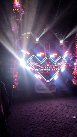The video shows a nighttime outdoor scene dominated by bright, colorful lights. In the center, a large, illuminated sign with red and white lettering, possibly a logo or brand name, is surrounded by a geometric shape outlined in white and blue lights. Multiple spotlights cast beams of light into the darkness, creating a dramatic atmosphere. To the left and right of the central sign, there are hints of decorative lighting structures, one appearing to be a curved, ornate display with pink and purple lights. The ground beneath is paved with what looks like brick or cobblestone. Silhouettes of people are visible in the background, and some are illuminated by the lights. One person in a light-colored garment stands near the central display, seemingly interacting with the equipment. The overall impression is of an event or performance with elaborate lighting effects. The video is shaky and the lighting is intense, sometimes causing overexposure.