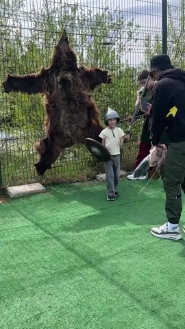 A boy dressed in a helmet and holding a shield and sword stands in front of a fence with a bear pelt hanging on it while three people take photos of him. The boy wears a light yellow shirt, gray pants, and black sneakers. He is standing on a green, artificial turf surface. Behind him is a chain-link fence, and pinned to the fence is a large, brown bear pelt. To the right of the boy, an individual in a green tunic and maroon pants stands, holding a sword. A person wearing a black hoodie and olive green pants kneels to take a photo with a smartphone, while another person in a black jacket stands nearby. There are several other helmets on the ground to the right of the boy. The scene appears to be a staged photo opportunity.