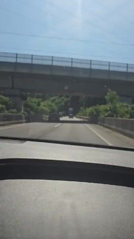 The video shows a first-person perspective from inside a vehicle driving on a highway. The dashboard is visible in the foreground. Ahead, the highway curves to the right, with a concrete barrier on the right side and a lush, green hillside on the left. Several cars are driving on the highway ahead. An overhead bridge crosses the highway in the distance. The sky is blue with bright sunlight creating glare on the windshield.