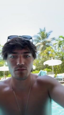 A young, shirtless man with brown hair and blue eyes stands in a swimming pool, partially submerged, holding the camera at eye level. He has sunglasses perched on his head and wears a silver necklace. The pool is filled with people, some wearing swimsuits and hats. Palm trees and white umbrellas are visible in the background, suggesting a resort setting. A large building can be seen further in the distance. The lighting is bright, indicating a sunny day. The man appears to be recording himself, possibly taking a selfie video.