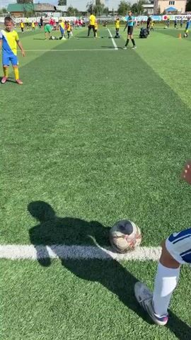 A short video clip shows a youth soccer practice or game. The perspective is from the side of the field, looking towards the action. Several young boys in yellow and blue uniforms are visible on the bright green artificial turf field. Some are running, some are kicking a soccer ball, and others are standing around. A few adults are also present, including what appears to be a referee in a yellow shirt and black shorts. The sky is blue with fluffy white clouds, and the sun is shining. The overall setting is a sports field with goals and fencing visible in the background.