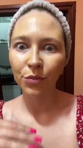 The media file is a close-up, selfie-style video of a woman with a light skin tone, wearing a white terrycloth headband. Her face is covered in a yellow mask, and she has wide, surprised-looking blue eyes. Her lips are pursed. She is wearing a red and white floral top. A gold hoop earring is visible in her right ear. Her left hand, with bright pink fingernails, is raised and touching her neck and collarbone area, with a ring on her ring finger. A small tattoo of a teacup is visible on her left shoulder. The background is dimly lit, with a wooden door frame and a hint of a kitchen or bathroom area. The lighting appears to be artificial, suggesting an indoor setting, possibly evening. The woman's expression and the setting suggest a personal grooming routine, possibly a face mask application. There is no visible text in the media file. The scene appears to be set in Podgorica, Montenegro, based on the provided context.