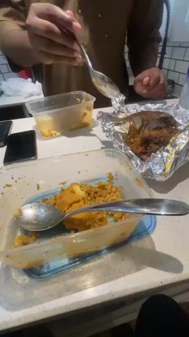 A medium, high-angle shot shows a close-up of a person serving themselves food. The person is an adult with dark skin and is wearing a tan shirt. Their left arm is visible as they hold a silver spoon in their right hand. The person uses the spoon to scoop meat out of a foil container on the right and places it onto a plastic food container on the left. The plastic food container contains rice and a yellow patty. The table is light in color and has two cell phones, one black and one dark, and another plastic food container with some food visible on it.