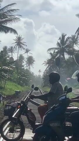 The video shows a view of railroad tracks in a tropical setting, likely Sri Lanka, given the location context. Palm trees line the tracks, and the sky is cloudy with patches of sunlight, suggesting it is daytime.

In the foreground, two people on a scooter are waiting near the railroad tracks. The scooter's front is visible, and both riders are wearing helmets. The person in the back is holding onto the person in the front. Further down the tracks, a second scooter with at least one rider is also visible.

The railroad tracks themselves are the central focus, curving into the distance and disappearing into a hazy, tropical landscape. The tracks appear to be in use, with ballast and ties visible. The overall impression is one of a rural or semi-rural area, possibly a railway crossing. The atmosphere seems calm, with the people on the scooter waiting patiently. The presence of multiple scooters suggests this might be a common mode of transport in the area. There is no visible text in the video.