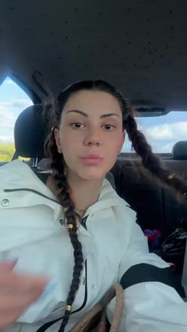 A young woman with dark hair in two braids with gold beads is sitting in the passenger seat of a car. She is wearing a white jacket. The sun is shining through the window, casting shadows on her face. She makes a silly face and then smiles.