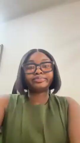 A person with dark hair and glasses smiles at the camera. The person wears a green sleeveless top. The person is indoors, and a wall is behind them. A black office chair is also visible behind them.